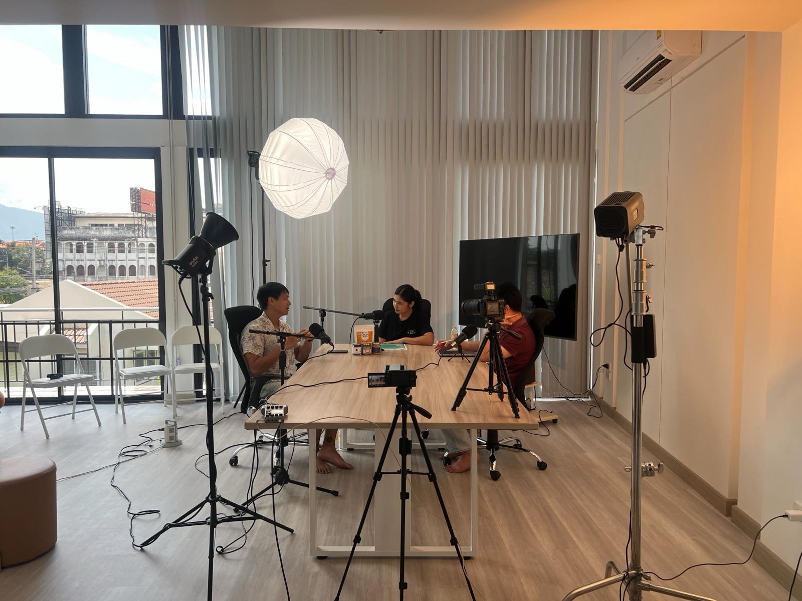 Three people recording around a table in the daylight studio, cameras on tripods and a balcony view behind